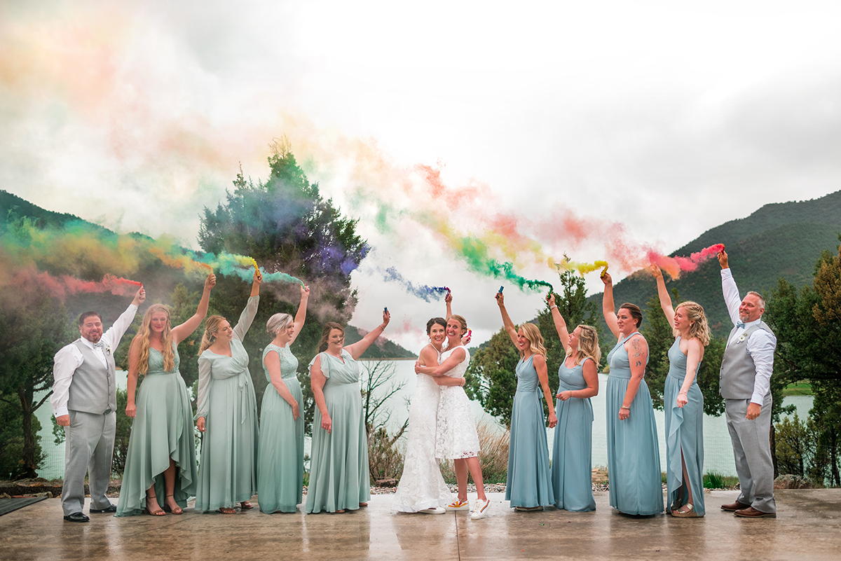 Two brides and the wedding party hold up smoke bombs in rainbow order